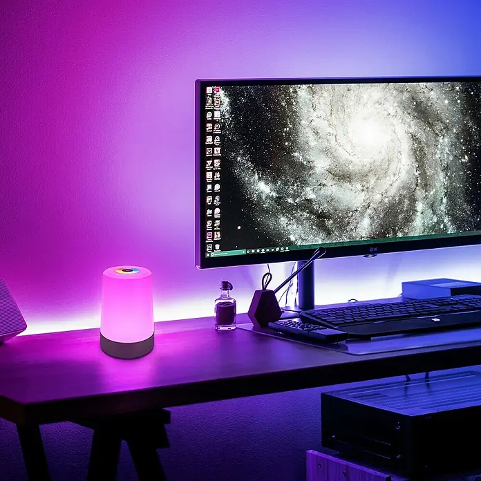 Brilo Akku-Tischleuchte Nubsi - Ø x H: 9 x 13 cm, Antharzit, RGBW, 2,8 W, IP44 - Ein RGB-Nachttischlampe mit Farbwechsel, die auf einem Schreibtisch neben einem Computer platziert ist. Erzeugt eine stimmungsvolle Beleuchtung.