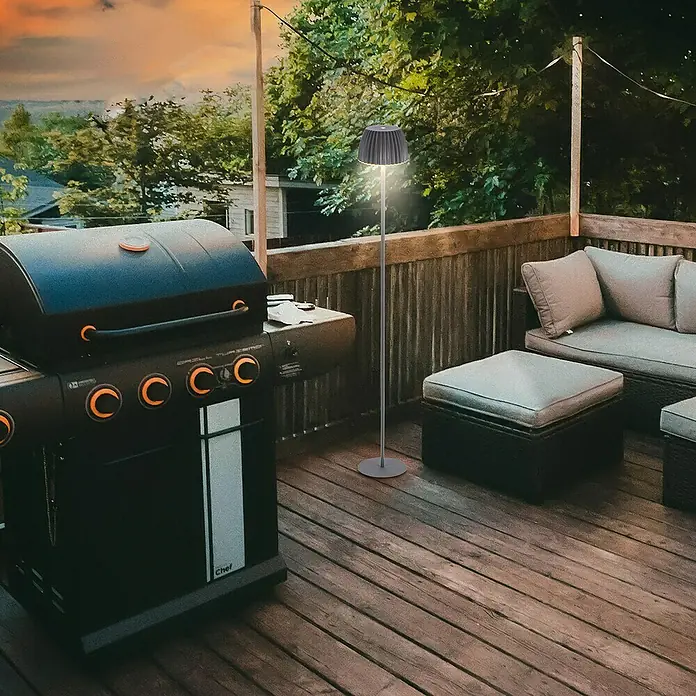 Brilo Riffle Akku-Stehleuchte Maxi - Ø x H: 19 x 124,1 cm, Anthrazit, Warmweiß, 4,2 W, IP44 - Aufnahme einer Stehleuchte im Freien mit Grill und Outdoor-Möbeln auf einer Holzterrasse, die eine entspannte Atmosphäre für Mahlzeiten im Freien bietet.