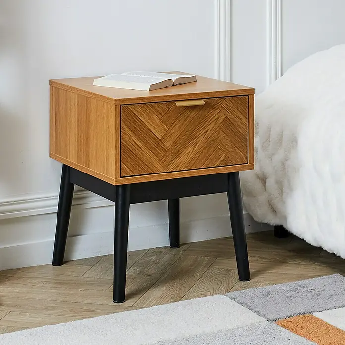 Home Deco Factory Felix Cómoda con mesitas - 3 pzs., Marrón/Negro, Acabado en espiga - Primer plano de una mesita de noche de madera con un cajón y un libro encima, creando un ambiente elegante en el dormitorio.
