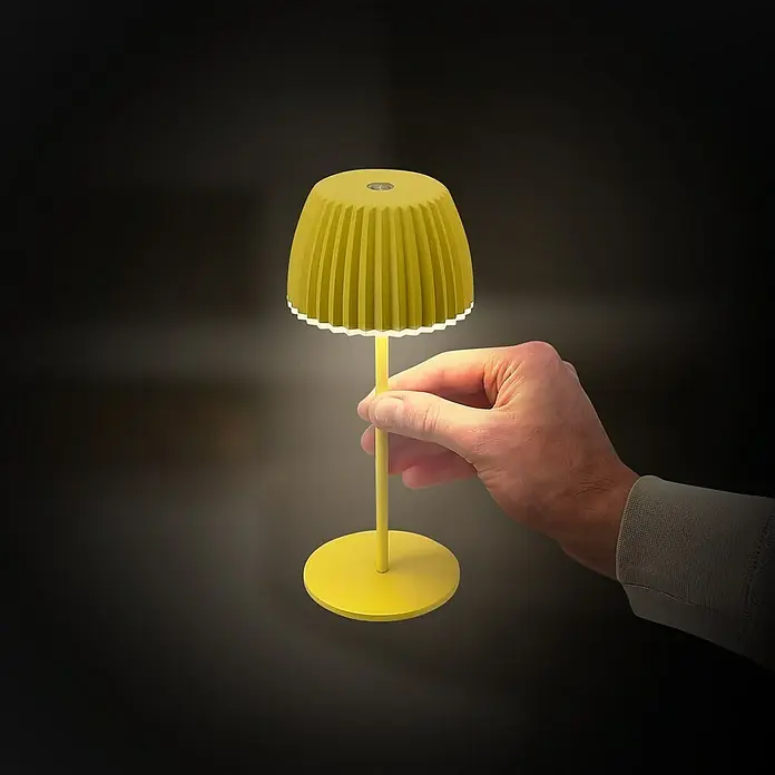 Brilo Riffle Akku-Tischleuchte - Ø x H: 8,5 x 20,1 cm, Gelb, Warmweiß, 1,5 W, IP44, Mini - Gelbe Tischleuchte wird von einer Hand gehalten, die ihr warmes Licht in einem dunklen Raum ausstrahlt.