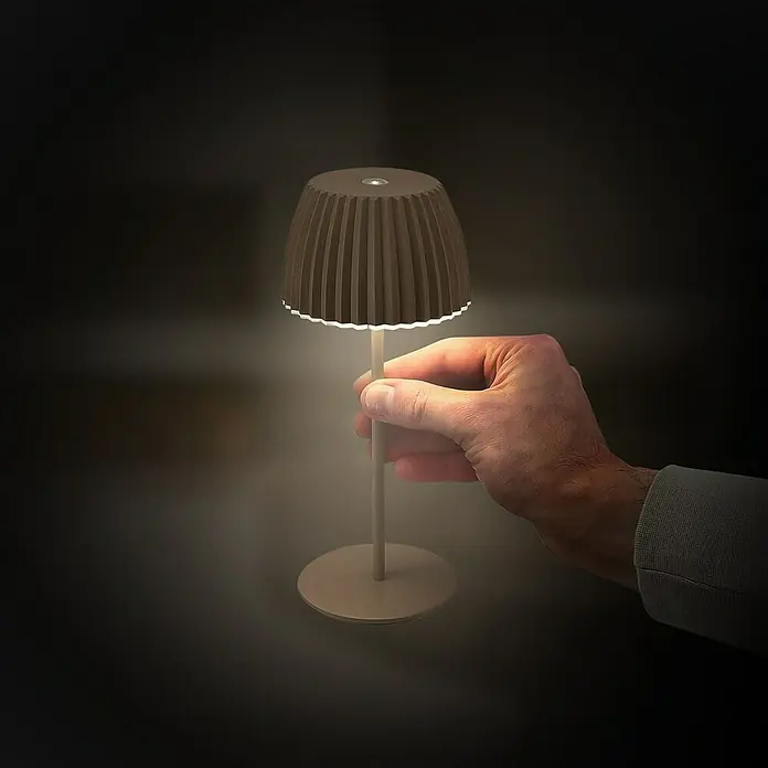 Brilo Riffle Akku-Tischleuchte - Ø x H: 8,5 x 20,1 cm, Braun, Warmweiß, 1,5 W, IP44, Mini - Eine Hand hält eine moderne, braune Tischleuchte mit plissiertem Schirm, die ein sanftes Licht ausstrahlt und eine gemütliche Atmosphäre erzeugt.