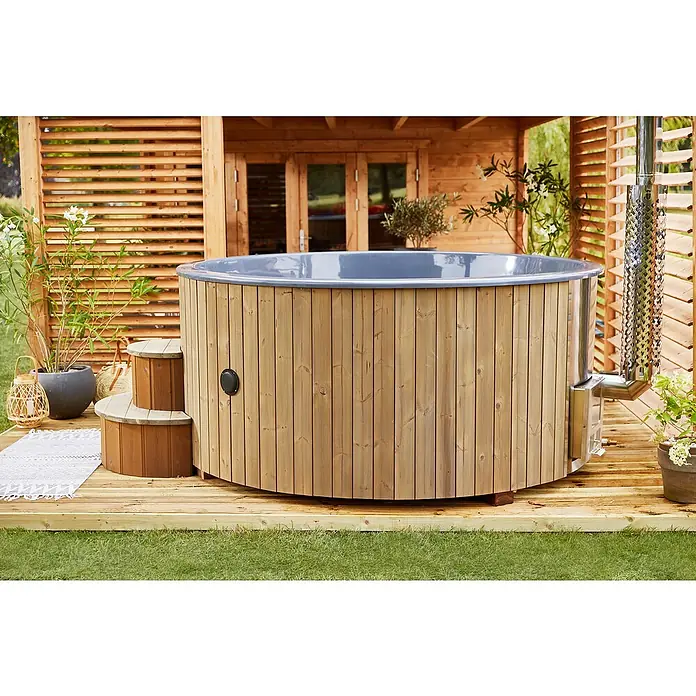 Tuindeco Hot Tub Deluxe - 200 cm, Kiefer, Natur, Max. Personenzahl: 6 Stk. - Holzbeheizter Badezuber mit Außentreppe und Edelstahlschornstein auf Holzterrasse vor einem Holzhaus.