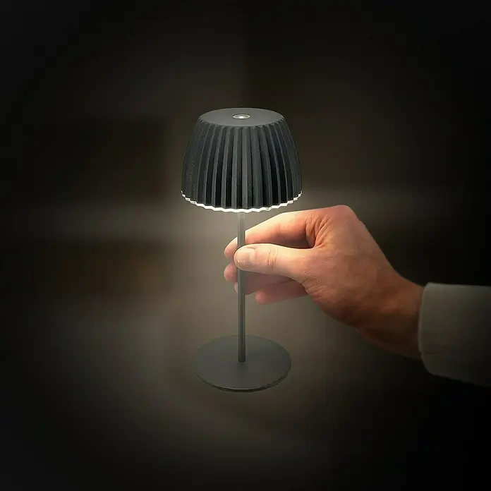 Brilo Riffle Akku-Tischleuchte - Ø x H: 8,5 x 20,1 cm, Anthrazit, Warmweiß, 1,5 W, IP44, Mini - Nahaufnahme einer stilvollen, kabellosen Tischleuchte in Anthrazit, die von einer Hand gehalten wird und sanftes Licht spendet.