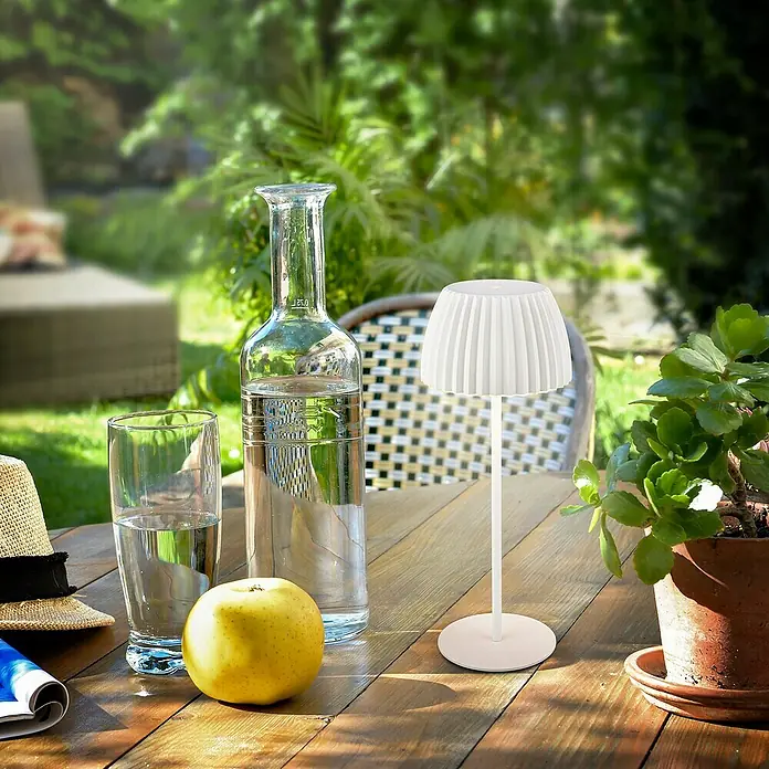 Brilo Riffle Akku-Tischleuchte - Ø x H: 14,3 x 36,4 cm, Weiß, Warmweiß, 2 W, IP44, Classic - Elegante weiße kabellose Lampe auf einem Holztisch im Freien, neben einem Glas Wasser, einer Flasche und einem Apfel für eine entspannte Atmosphäre.