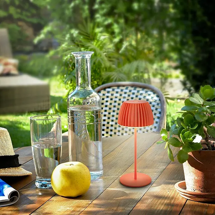 Brilo Riffle Akku-Tischleuchte - Ø x H: 8,5 x 20,1 cm, Orange, Warmweiß, 1,5 W, IP44, Mini - Nahaufnahme einer warmen, orangefarbenen tragbaren Lampe auf einem Holztisch im Freien.
