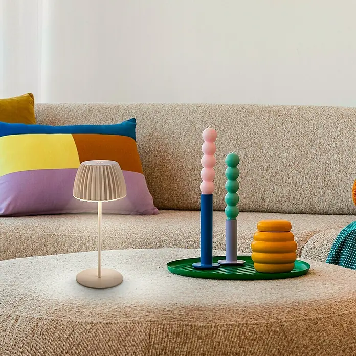 Brilo Riffle Akku-Tischleuchte - Ø x H: 8,5 x 20,1 cm, Beige, Warmweiß, 1,5 W, IP44, Mini - Tischleuchte auf einem Tisch neben bunten Kerzen und Kissen auf einem Sofa.