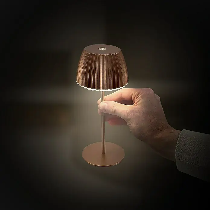 Brilo Riffle Akku-Tischleuchte - Ø x H: 8,5 x 20,1 cm, Kupfer, Warmweiß, 1,5 W, IP44, Mini - Nahaufnahme einer Hand, die eine dekorative, dimmbare LED-Tischleuchte mit plissiertem Schirm und warmem Licht hält.