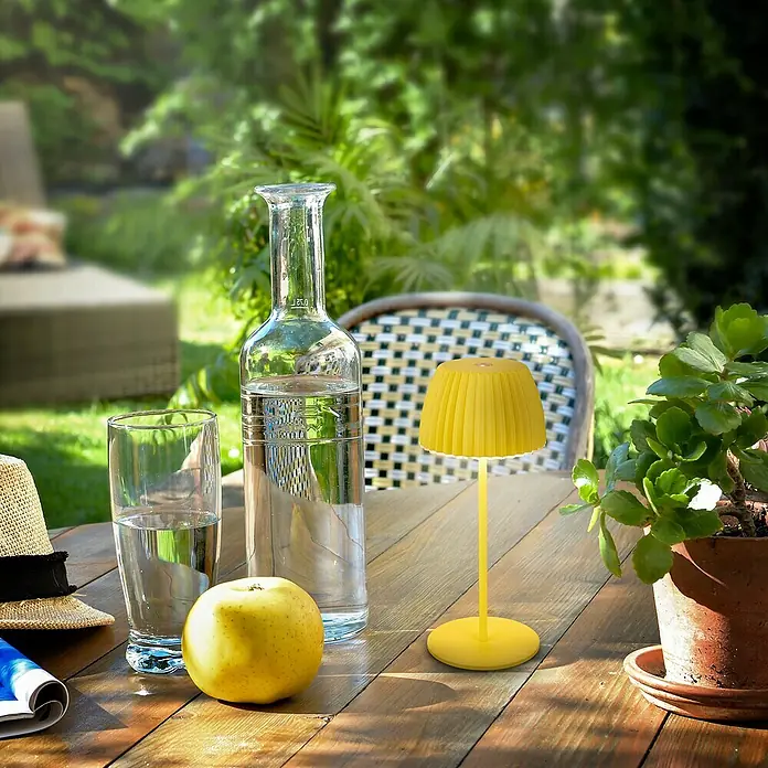 Brilo Riffle Akku-Tischleuchte - Ø x H: 8,5 x 20,1 cm, Gelb, Warmweiß, 1,5 W, IP44, Mini - Gartentisch mit gelber Lampe, Wasserflasche und Apfel.