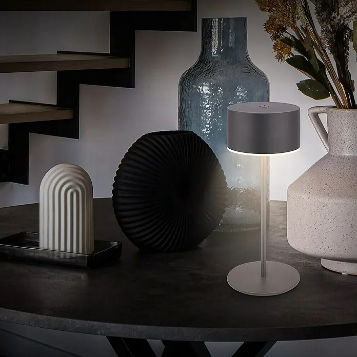 Brilo Akku-Tischleuchte Kira - Ø x H: 9 x 18 cm, Anthrazit, Warmweiß, 1,5 W, IP44 - Elegante Tischleuchte in Grau auf einem Tisch mit dekorativen Objekten. Schafft eine stimmungsvolle Atmosphäre.