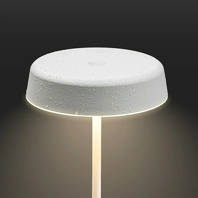 Brilo Akku-Tischleuchte Pala - Ø x H: 13 x 36,5 cm, Altweiß, Warmweiß, 2,6 W, IP44 - Das Bild zeigt eine moderne, weiße Lampe mit einem runden Lampenschirm, der mit Wassertropfen bedeckt ist, vor einem dunklen Hintergrund.