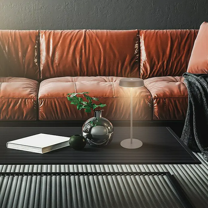 Brilo Akku-Tischleuchte Pala - Ø x H: 13 x 36,5 cm, Anthrazit, Warmweiß, 2,6 W, IP44 - Nahaufnahme einer modernen Tischleuchte auf einem Tisch mit Buch, Vase und Dekoration vor einem Ledersofa.