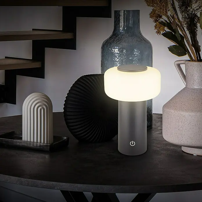 Brilo Akku-Tischleuchte Juna - Ø x H: 13 x 20,5 cm, Anthrazit, Warmweiß, 1,8 W, IP44 - Dekorativer Tisch mit einer modernen Lampe, einer dunklen Vase und einer hellen Skulptur. Die Lampe spendet warmes Licht.