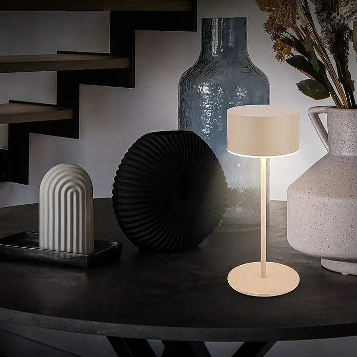 Brilo Akku-Tischleuchte Kira - Ø x H: 9 x 18 cm, Beige, Warmweiß, 1,5 W, IP44 - Elegante Tischleuchte mit rundem Lampenschirm, umgeben von dekorativen Vasen und Objekten.