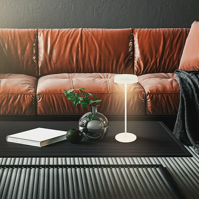 Brilo Akku-Tischleuchte Pala - Ø x H: 13 x 36,5 cm, Altweiß, Warmweiß, 2,6 W, IP44 - Nahaufnahme einer modernen LED-Tischleuchte auf einem Tisch, daneben ein Buch, eine grüne Kugel und eine Vase mit Pflanzen.