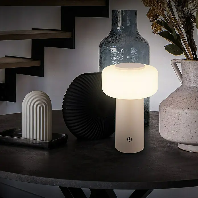 Brilo Akku-Tischleuchte Juna - Ø x H: 13 x 20,5 cm, Beige, Warmweiß, 1,8 W, IP44 - Nahaufnahme einer modernen Tischleuchte, die ein warmes Licht ausstrahlt, umgeben von dekorativen Objekten auf einem eleganten Tisch.