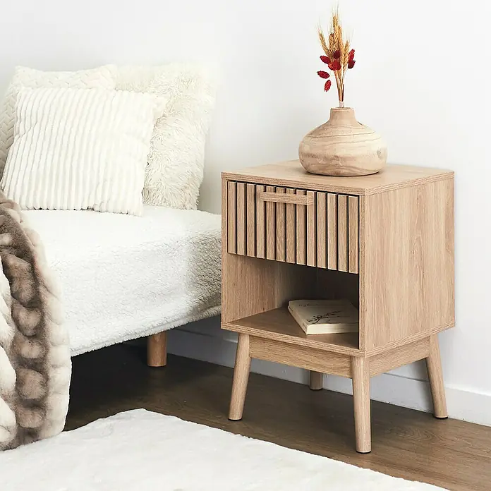 Home Deco Factory Klaus Cómoda con mesitas - 3 pzs., Marrón, Diseño acanalado - La imagen muestra una mesita de noche de madera clara con un cajón y un estante abierto donde descansa un libro. Un jarrón adorna la parte superior.