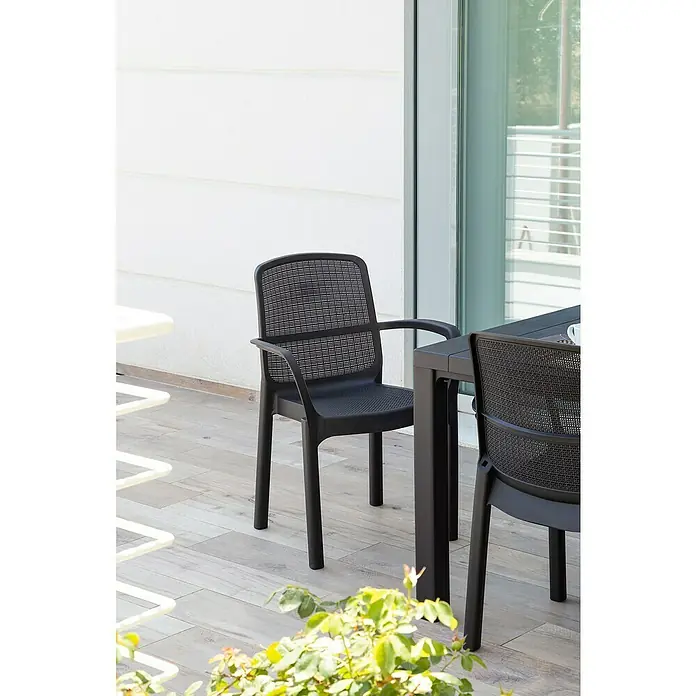 Gartensessel Sirolo - Schwarz, Kunststoff - Schwarzer Armlehnstuhl aus Kunststoff mit Netzmuster, ideal für den Außenbereich, auf einer Terrasse neben einem Tisch.