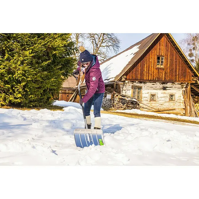 Freund Victoria Schneeschieber Profi - Breite: 50 cm, Langstiel, Material Stiel: Aluminium - Frau schippt mit einer blauen Schneeschaufel Schnee vor einem Holzhaus. Die Schaufel ist robust und ideal für winterliche Bedingungen.
