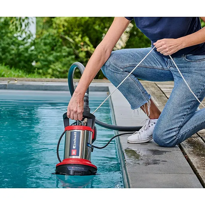 Einhell Bomba para aguas residuales Flutset GE-DP 7535 - Capacidad de bombeo máx.: 18.500 l/h, Profundidad de inmersión: 7 m - Una mujer introduce una bomba de agua sucia Einhell roja en una piscina. La bomba está conectada a una manguera y un cable.