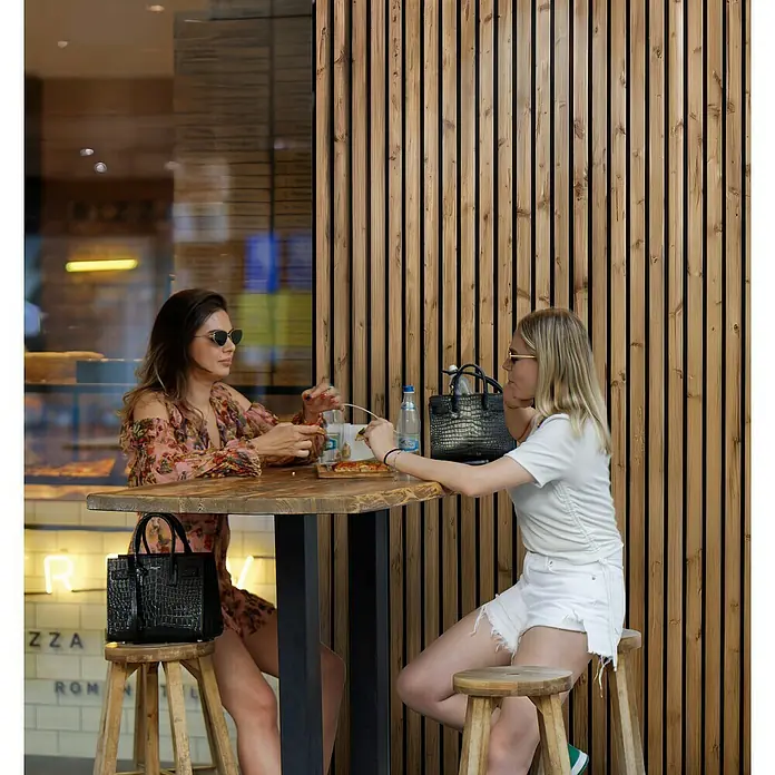 Akustikpaneel Outdoor HydroLine - Braun, 2.400 x 561 x 28 mm - Zwei Frauen sitzen an einem Tisch mit Handtaschen. Die schwarze Handtasche links ist die Saint Laurent Sac De Jour Nano.
