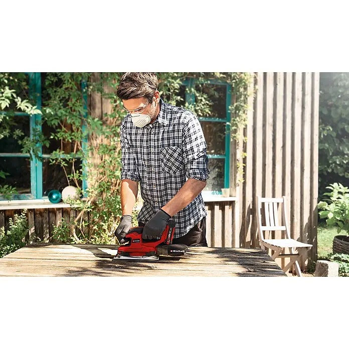Einhell Power X-Change 18V Akku-Schwingschleifer - 18 V, Ohne Akku, 14.000 U/min - 24.000 U/min - Ein Mann schleift eine Holzplatte mit einem roten Einhell-Schleifgerät, während er Schutzbrille und Maske trägt.