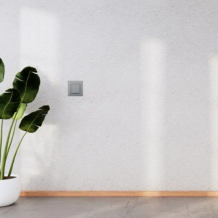 Legrand Valena Next with Netatmo Conmutador On/Off - Aluminio, Mate, Montaje en la pared - Primer plano de una pared de cemento gris con un interruptor de luz empotrado y una planta en maceta. La luz difusa enfatiza la textura.