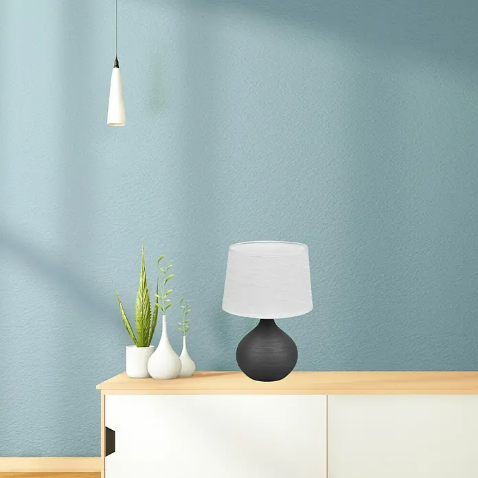 Trio Lighting Lámpara de sobremesa Martin - 40 W, L x An x Al: 20 x 20 x 29 cm, Negro, E14 - Lámpara de mesa decorativa con pantalla blanca y base oscura, sobre una mesa clara frente a una pared azul verdosa.