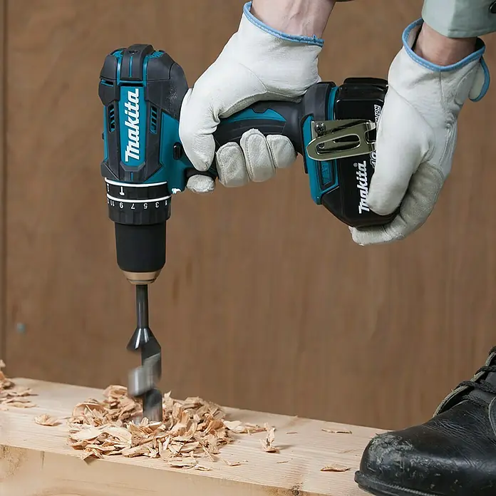 Makita LXT 18V Akku-Schlagbohrschrauber DHP482RFX9 - 18 V, 2 Akkus, 3 Ah - Makita Akku-Bohrschrauber beim Bohren in Holz, mit Spänen um das Bohrloch. Schutzhandschuhe und Arbeitsschuh sind sichtbar.