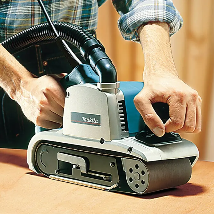 Makita Elektro-Bandschleifmaschine 9403J - 1.200 W - Ein Makita Bandschleifer wird von einer Person auf einer Holzoberfläche bedient, während ein Staubsaugerschlauch befestigt ist.