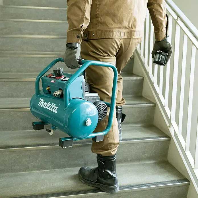 Makita XGT 40V max. Akku-Kompressor AC001GZ - 40 V, Ohne Akku, 9,3 bar - Ein Mann trägt einen tragbaren Makita-Luftkompressor eine Treppe hinauf, ideal für Baustellen.