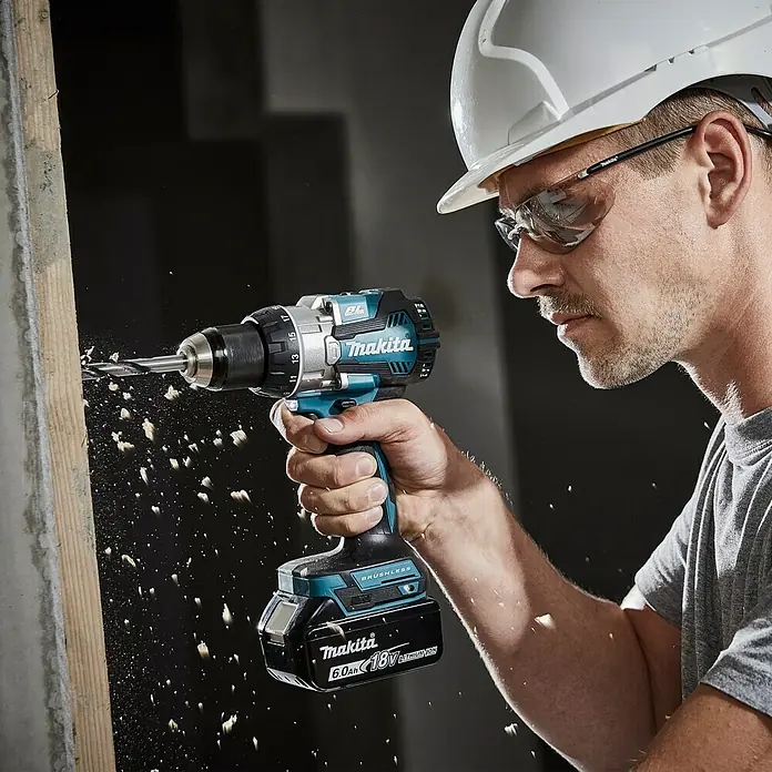 Makita LXT 18V Akku-Schlagbohrschrauber DHP489RTJ - 18 V, 2 Akkus, 5 Ah, 73 Nm - Der Makita Akku-Bohrschrauber beim Bohren in Holz, mit Spänen die herumfliegen. Der Handwerker trägt Schutzbrille und Helm.