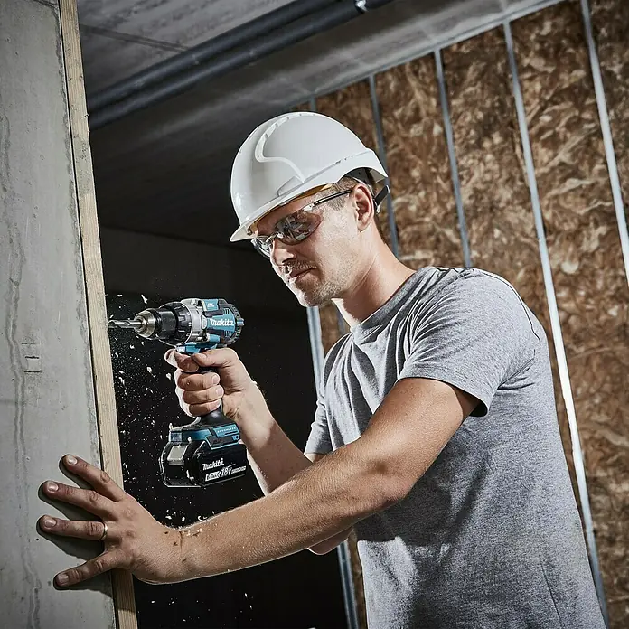 Makita LXT 18V Akku-Schlagbohrschrauber DHP489RTJ - 18 V, 2 Akkus, 5 Ah, 73 Nm - Ein Bauarbeiter bohrt mit einem Makita Akku-Bohrschrauber ein Loch in eine Wand. Schwebende Holzspäne.