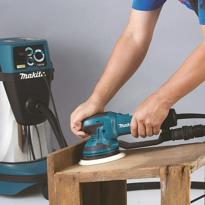Makita Elektro-Exzenterschleifer BO6050J - 750 W, Durchmesser Schleifteller: 150 mm - Nahaufnahme eines Makita Exzenterschleifers beim Schleifen von Holz, angeschlossen an einen Staubsauger für Staubabsaugung.