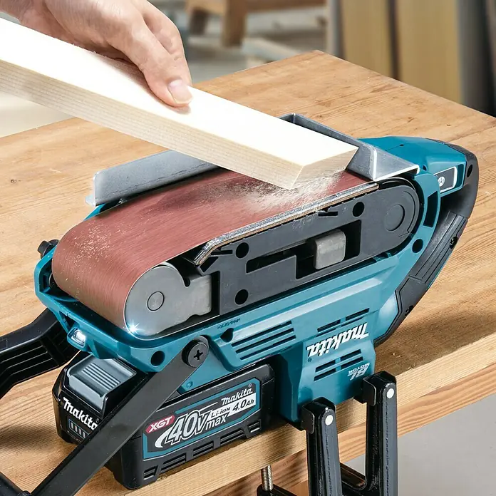 Makita XGT 40V max. Akku-Bandschleifer BS001GZ - 40 V, Ohne Akku - Nahaufnahme eines Makita Akku-Bandschleifers in Aktion, der ein Holzstück bearbeitet. Das Werkzeug ist auf einer Werkbank befestigt.