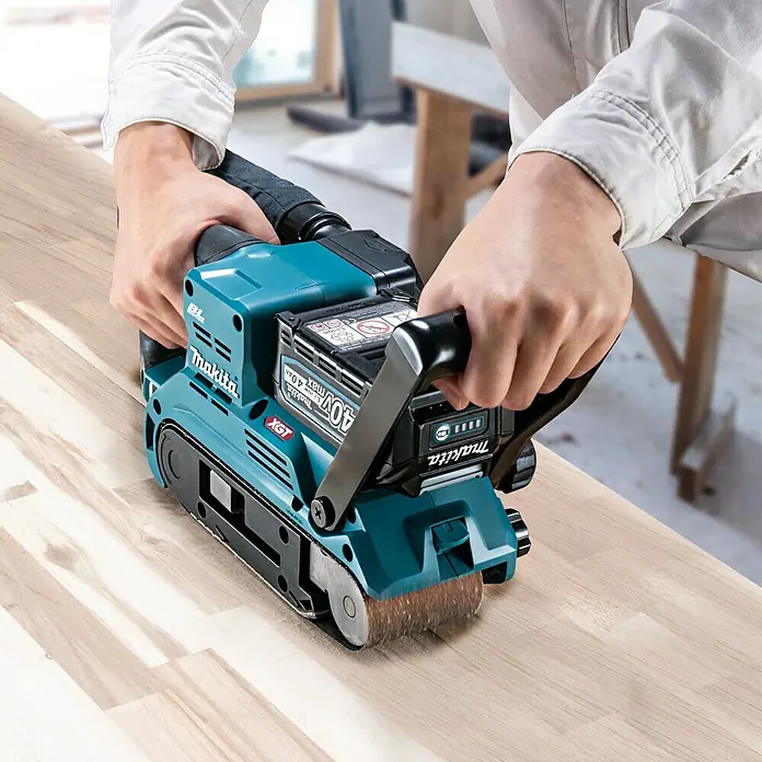 Makita XGT 40V max. Akku-Bandschleifer BS001GZ - 40 V, Ohne Akku - Ein Makita Akku-Bandschleifer XGT wird auf einem Holzbrett von einer Person mit beiden Händen bedient.