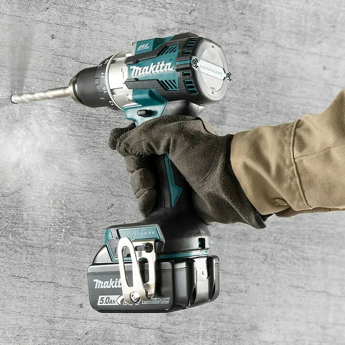 Makita LXT 18V Akku-Schlagbohrschrauber DHP489RTJ - 18 V, 2 Akkus, 5 Ah, 73 Nm - Nahaufnahme eines Makita Akku-Bohrschraubers in Aktion, der in eine graue Wand bohrt, mit Staubpartikeln in der Luft.