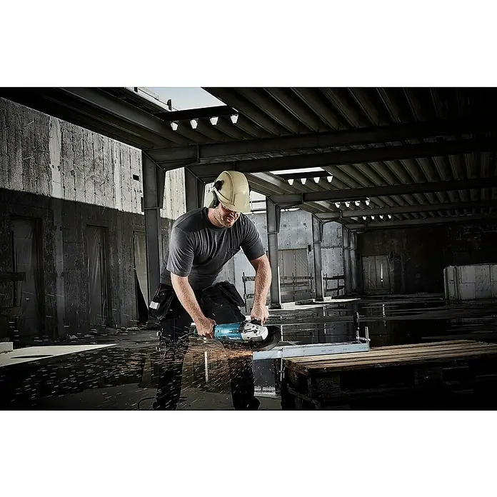 Makita Elektro-Winkelschleifer GA 9020 RF - 2.200 W, Durchmesser Scheibe: 230 mm, Leerlaufdrehzahl: 6.600 U/min - Ein Bauarbeiter mit Schutzhelm benutzt einen Winkelschleifer in einer Lagerhalle, um Metall zu schneiden.
