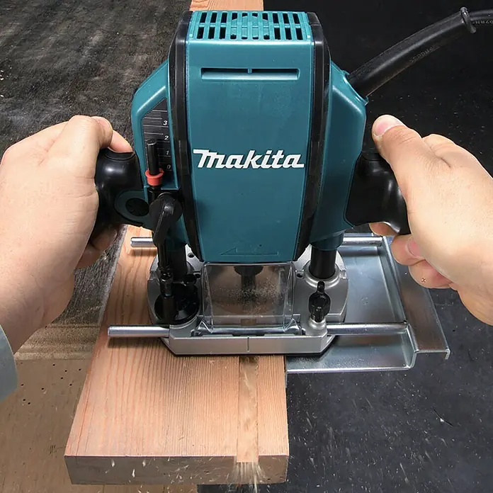 Makita Oberfräse RP0900J - 900 W, 0 U/min - 27.000 U/min, Frästiefe: 35 mm - Nahaufnahme einer Makita Oberfräse in Aktion, die ein Holzstück bearbeitet. Das Werkzeug wird von zwei Händen gehalten.