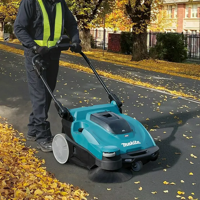 Makita XGT 40V max. Akku-Handkehrmaschine VS001GZ - 40 V, Arbeitsbreite: 650 mm, 24 l - Ein Mann bedient eine blaue Makita Kehrmaschine auf einer mit Blättern bedeckten Straße.