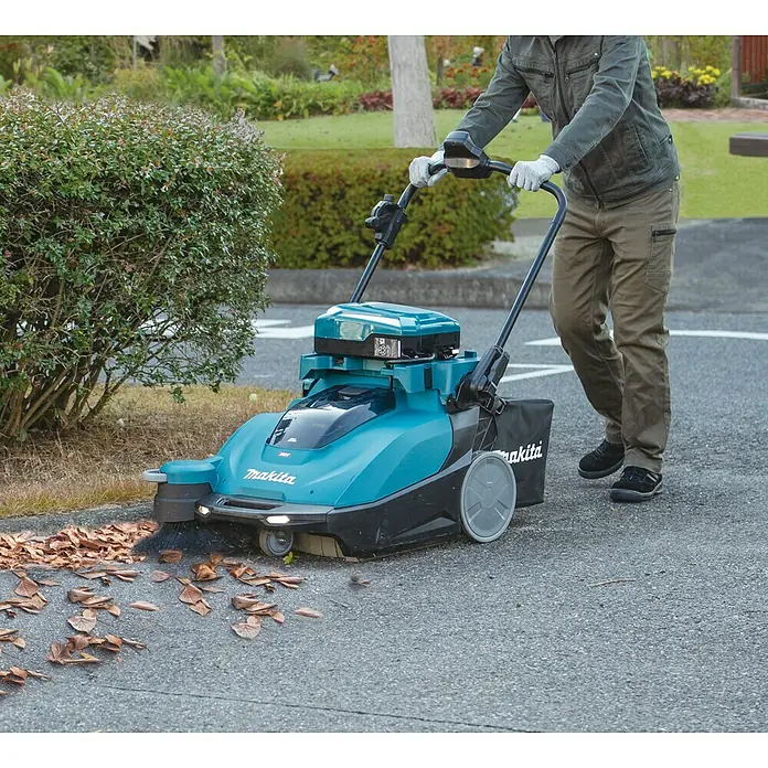 Makita XGT 40V max. Akku-Handkehrmaschine VS001GZ - 40 V, Arbeitsbreite: 650 mm, 24 l - Ein Mann benutzt eine Makita Kehrmaschine im Freien, um Laub zu entfernen. Das Gerät ist blaugrün mit schwarzen Akzenten.
