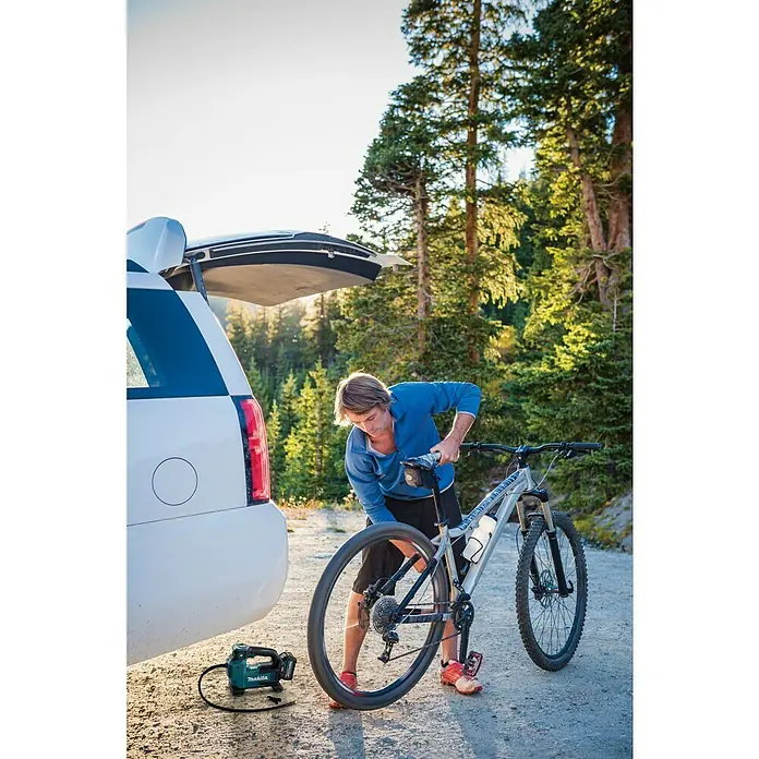 Makita XGT 40V max. Akku-Kompressor MP001GZ - 40 V, Ohne Akku, 11,1 bar - Ein Mann repariert sein Fahrrad neben einem Auto mit geöffnetem Kofferraum. Im Vordergrund ist ein Luftkompressor von Makita zu sehen.