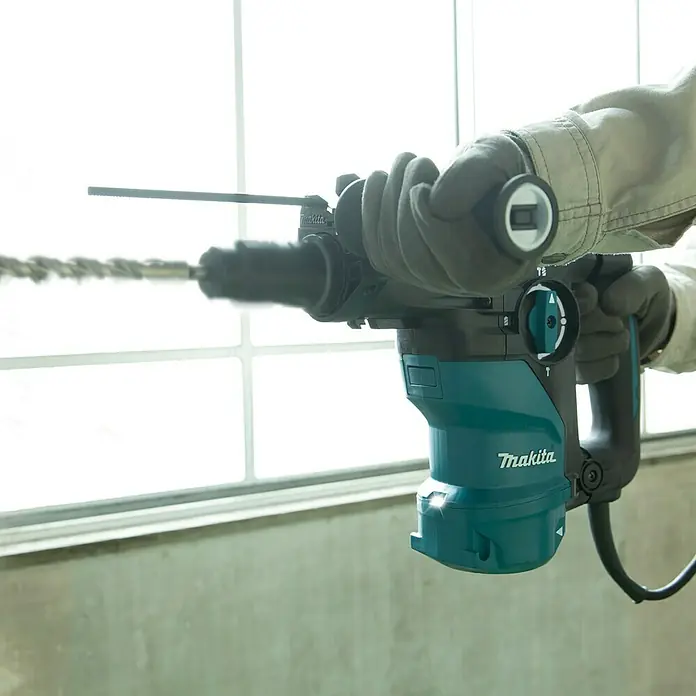 Makita Kombihammer HR3012FCJ - 1.050 W, 3,9 J, SDS-Plus-Aufnahme - Nahaufnahme einer Makita-Bohrmaschine mit Bohrer, die von einer Person mit Handschuhen bedient wird.