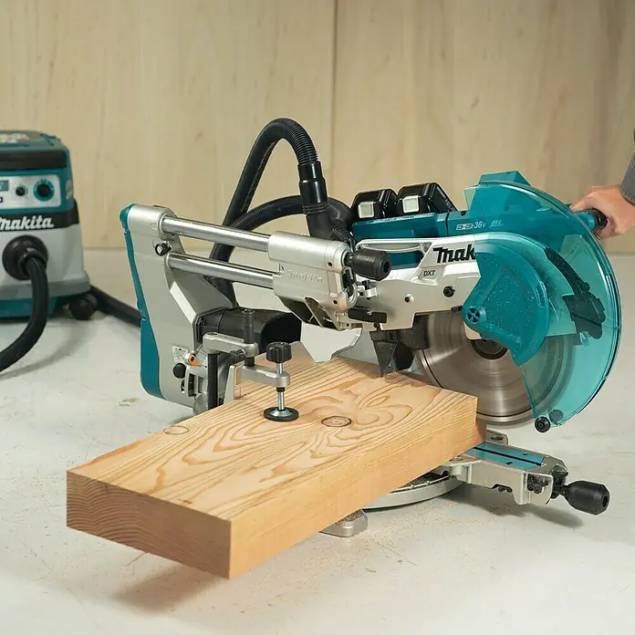 Makita LXT 18V Akku-Kapp- & Gehrungssäge DLS211ZU - 36 V, Ohne Akku - Aufnahme einer Makita-Kapp- und Gehrungssäge mit einem Holzstück, das mit einer Klemme befestigt ist, und einem Staubsauger im Hintergrund.