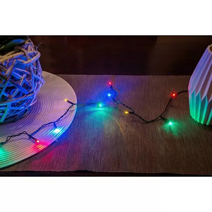 Konstsmide LED-Lichterkette Micro LED - Innen, Lichtfarbe: Mehrfarbig, Anzahl Leuchtmittel: 100 Stk., Netzbetrieben - Bunte Lichterkette mit mehreren LEDs, die einen festlichen Glanz auf eine strukturierte Tischdecke zwischen Deko-Objekten wirft.