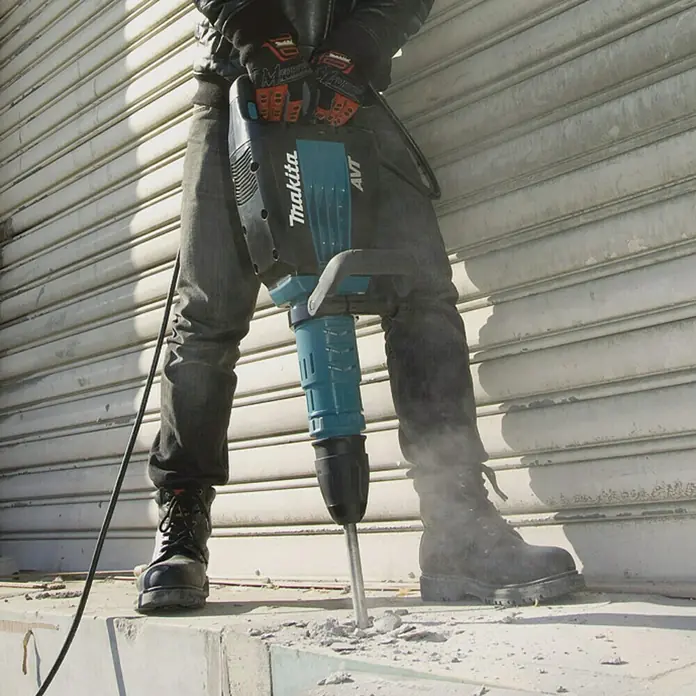 Makita Abbruchhammer HM1214C - 1.510 W, 19,9 J, SDS-Max-Aufnahme - Ein Bauarbeiter benutzt einen Makita-Bohrhammer zum Aufbrechen von Beton, wobei Staub aufwirbelt.