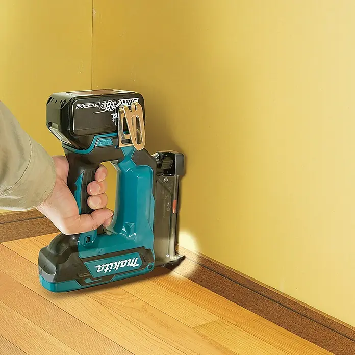 Makita LXT 18V Akku-Nagler DPT353Z - 18 V, Ohne Akku, Länge Nagel: 15 mm - 35 mm - Eine Makita Akku-Nagelpistole wird an einer Fußleiste auf einem Holzboden verwendet. Das Gerät ist blaugrün und schwarz mit einem Akku oben.