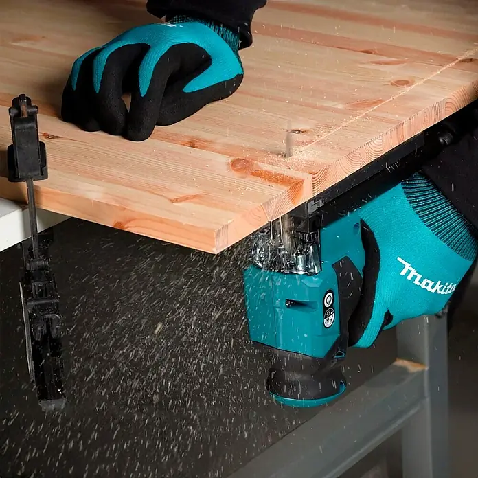 Makita XGT 40V max. Akku-Pendelhubstichsäge JV001GZ - 40 V, Ohne Akku, Leerlaufhubzahl: 800 /min - 3.500 /min - Eine Makita-Säge schneidet durch ein Holzbrett, Späne fliegen. Der Bediener trägt Schutzhandschuhe. Eine Zwinge ist auch im Bild.