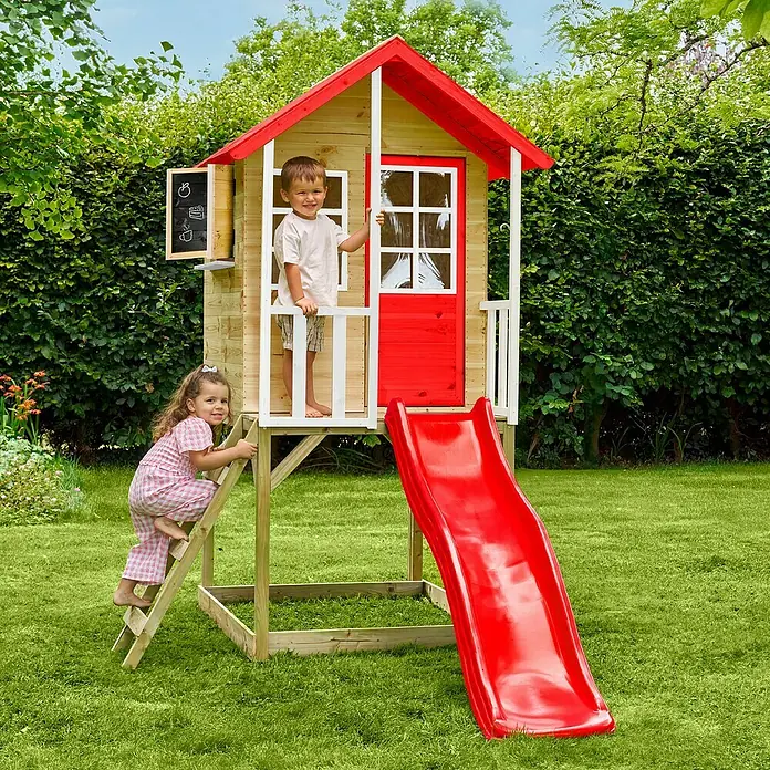 Zwerkules Spielhaus Lolla - L x B x H: 270 x 148 x 224 cm, Holz, Natur/Rot, Mit Stelzen & Rutsche - Ein hölzernes Spielhaus mit Rutsche und Leiter im Garten, in dem ein Junge steht und ein Mädchen die Leiter hochklettert.