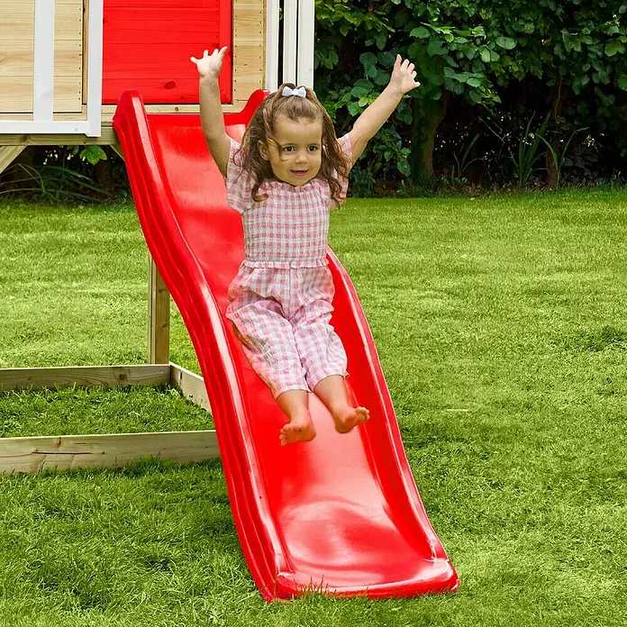 Zwerkules Spielhaus Lolla - L x B x H: 270 x 148 x 224 cm, Holz, Natur/Rot, Mit Stelzen & Rutsche - Mädchen saust mit einem roten Kinderspielplatzrutsche hinunter, die sich perfekt zum Spielen im Garten eignet.