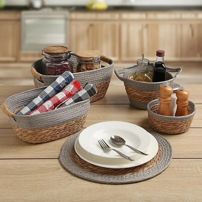 Versa Cesta Gris - L x An x Al: 27 x 20 x 12 cm, Gris - Cinco cestas de junco tejidas con diferentes contenidos están dispuestas sobre una mesa, con ajustes de lugar sobre un posavasos tejido a juego en primer plano.